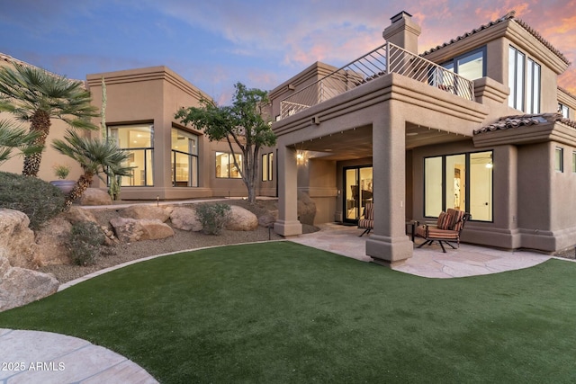 back house at dusk featuring a patio, a balcony, and a lawn