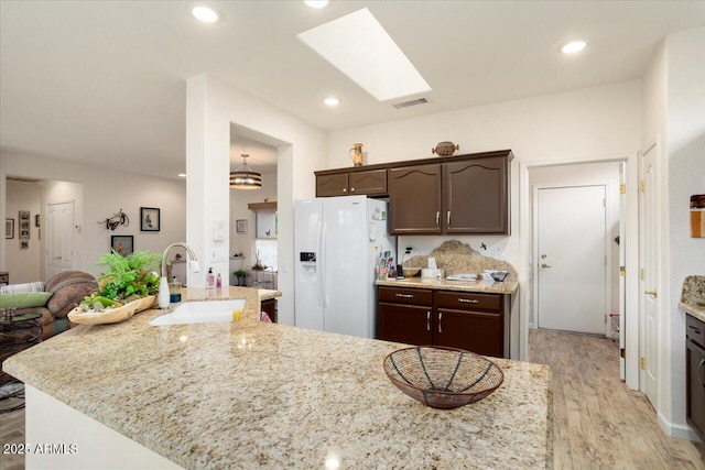 kitchen with dark brown cabinetry, sink, light stone counters, a skylight, and white refrigerator with ice dispenser