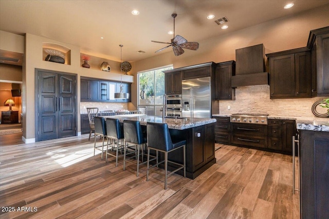 kitchen with appliances with stainless steel finishes, backsplash, custom exhaust hood, pendant lighting, and a center island with sink