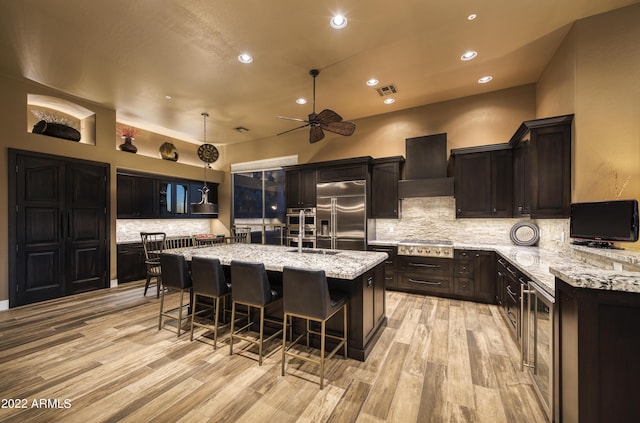 kitchen with stainless steel appliances, decorative light fixtures, a kitchen island with sink, custom range hood, and light wood-type flooring