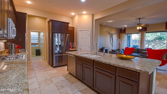 kitchen featuring stainless steel appliances, a center island with sink, sink, light stone counters, and ceiling fan