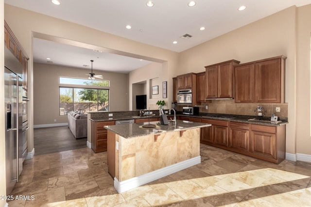 kitchen with appliances with stainless steel finishes, a kitchen island with sink, hanging light fixtures, dark stone countertops, and decorative backsplash