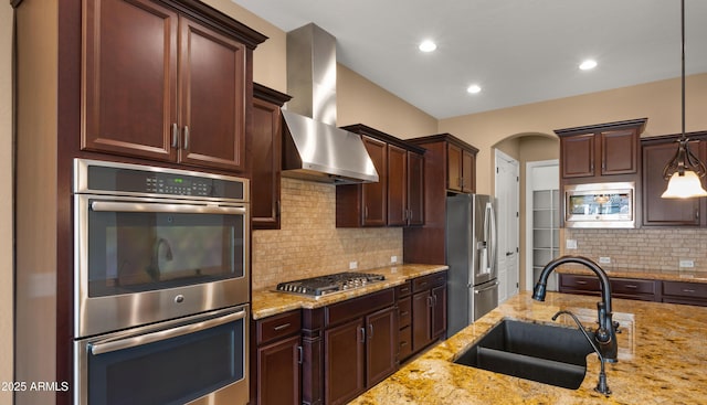 kitchen featuring light stone countertops, a sink, stainless steel appliances, wall chimney range hood, and backsplash