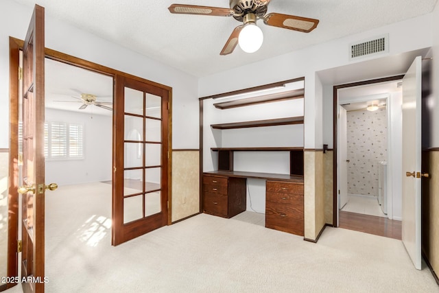 unfurnished bedroom featuring built in desk, ceiling fan, light carpet, a textured ceiling, and french doors