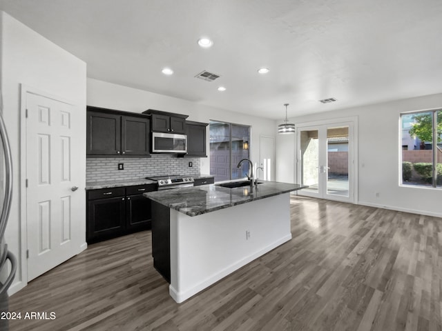 kitchen with dark wood-type flooring, sink, stainless steel appliances, a center island with sink, and dark stone countertops