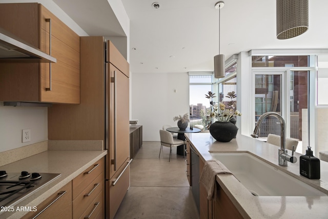 kitchen with a sink, stainless steel gas cooktop, range hood, brown cabinets, and built in fridge