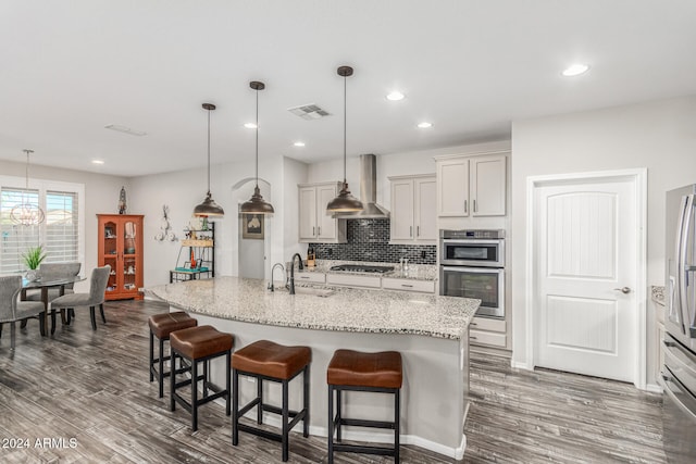 kitchen featuring wall chimney exhaust hood, decorative light fixtures, an island with sink, and light stone countertops