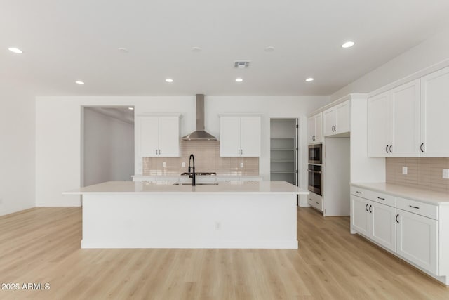 kitchen with wall chimney exhaust hood, an island with sink, light countertops, and white cabinetry