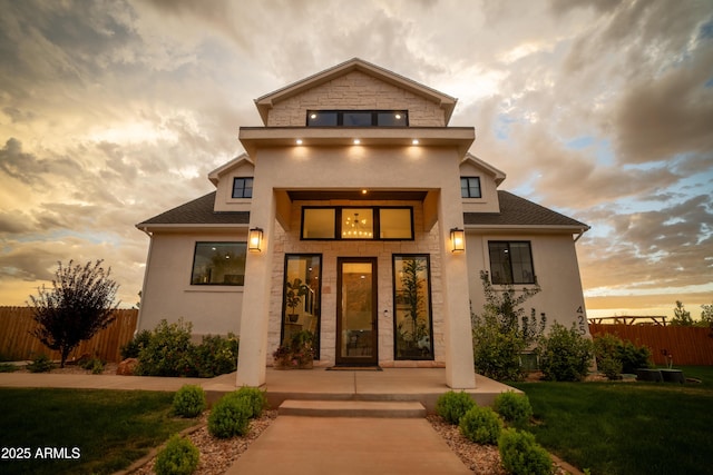 exterior entry at dusk with stone siding, a shingled roof, and fence