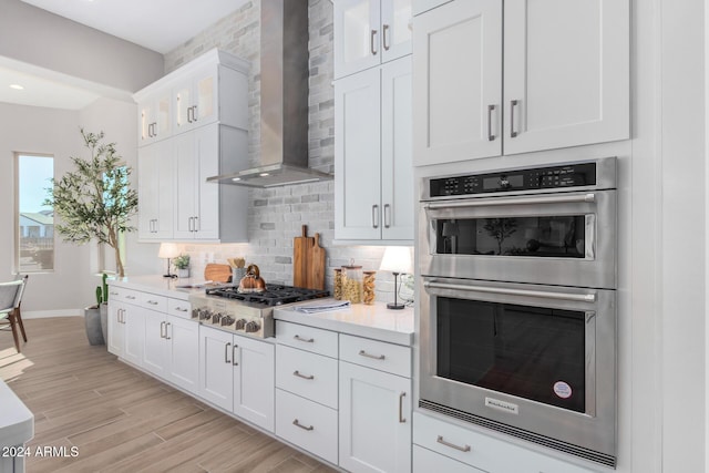 kitchen featuring wall chimney exhaust hood, white cabinetry, tasteful backsplash, light wood-type flooring, and appliances with stainless steel finishes