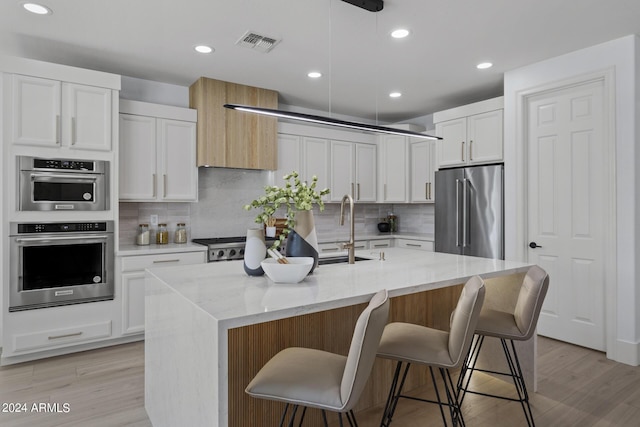 kitchen featuring a kitchen island with sink, appliances with stainless steel finishes, decorative light fixtures, light stone counters, and white cabinetry