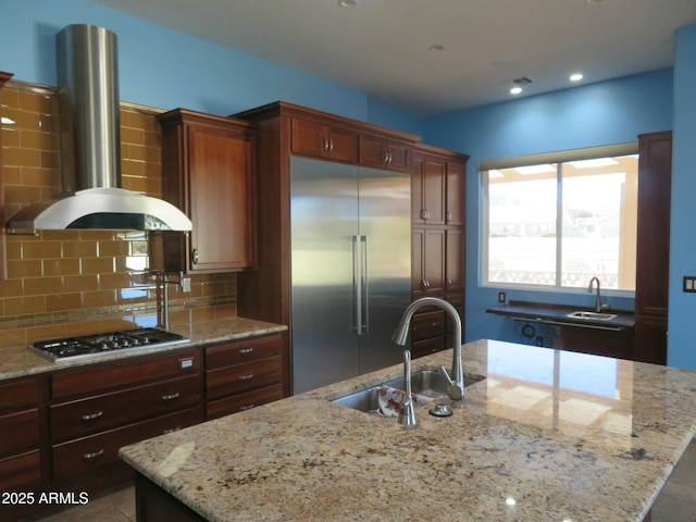 kitchen with wall chimney range hood, stainless steel appliances, an island with sink, and a sink
