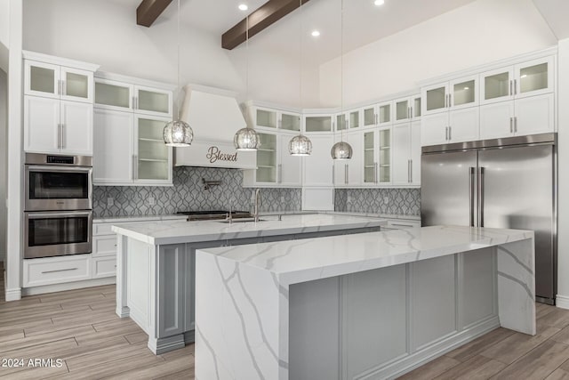 kitchen featuring appliances with stainless steel finishes, light stone counters, a large island with sink, beam ceiling, and a high ceiling