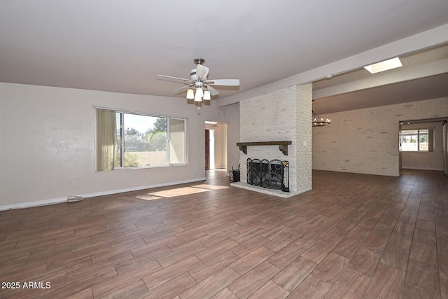 unfurnished living room with ceiling fan with notable chandelier, wood finished floors, brick wall, baseboards, and a brick fireplace