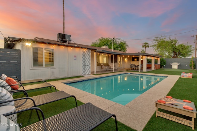 pool at dusk featuring a lawn, cooling unit, and a patio