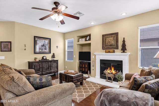 living area with visible vents, ceiling fan, built in features, dark wood-style floors, and a glass covered fireplace