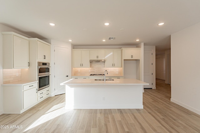 kitchen with white cabinetry, a kitchen island with sink, stainless steel double oven, and light hardwood / wood-style floors