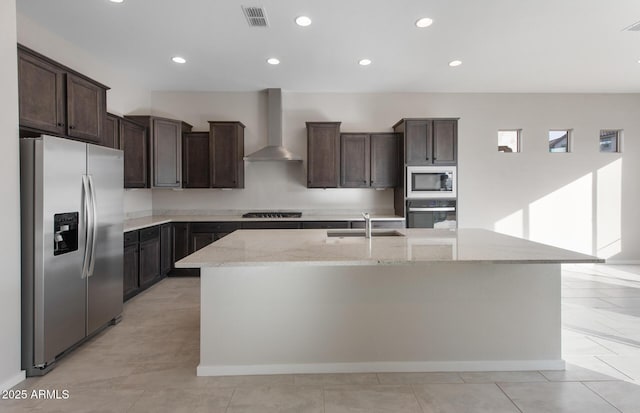 kitchen featuring light stone countertops, stainless steel appliances, a kitchen island with sink, sink, and wall chimney range hood