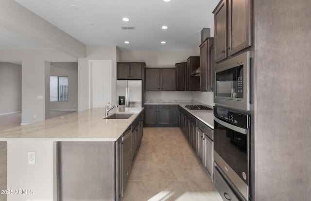 kitchen featuring a center island with sink, sink, light stone countertops, dark brown cabinets, and stainless steel appliances