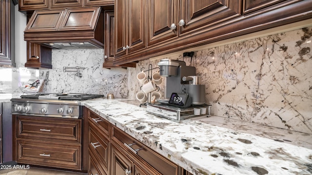 kitchen with tasteful backsplash, dark brown cabinetry, light stone countertops, stainless steel gas cooktop, and custom exhaust hood