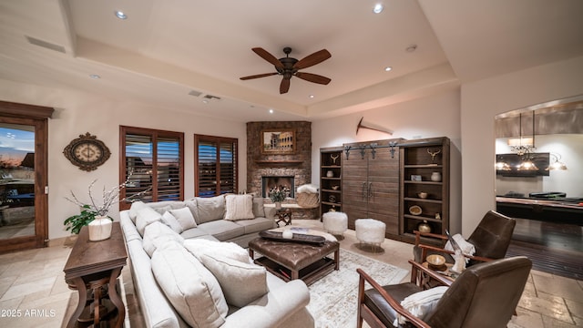 living room featuring ceiling fan, a tray ceiling, and a stone fireplace