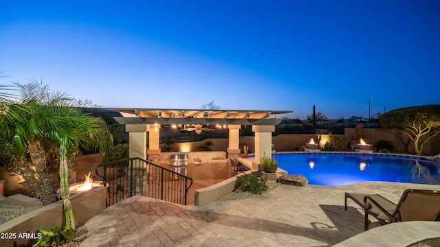 pool at dusk featuring exterior kitchen, a pergola, a patio area, and an outdoor fire pit