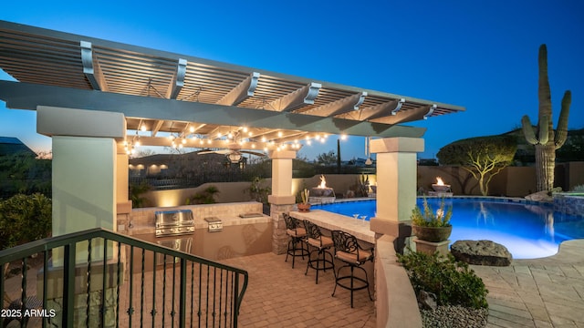 pool at night with a patio, a bar, a grill, exterior kitchen, and a pergola