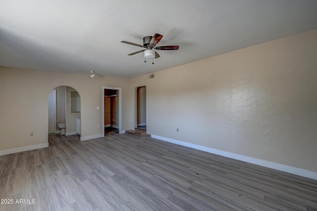 empty room featuring arched walkways, a ceiling fan, light wood-style floors, and baseboards
