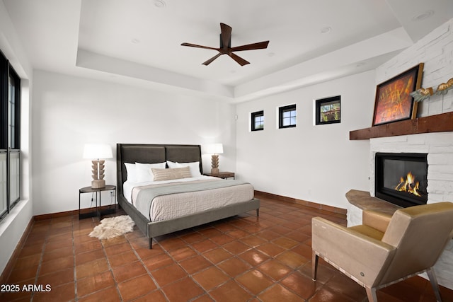 bedroom featuring dark tile patterned floors, a raised ceiling, ceiling fan, and a stone fireplace