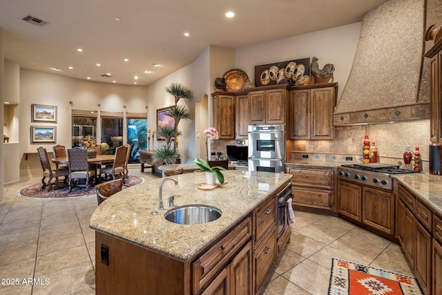 kitchen featuring appliances with stainless steel finishes, a sink, an island with sink, decorative backsplash, and light tile patterned floors