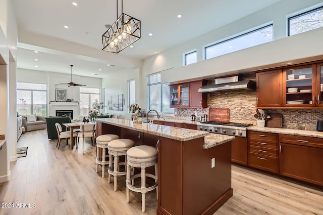 kitchen with light stone countertops, wall chimney range hood, an island with sink, decorative light fixtures, and stainless steel gas stovetop