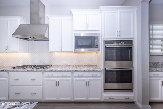 kitchen with wall chimney range hood, appliances with stainless steel finishes, tasteful backsplash, light stone counters, and white cabinetry