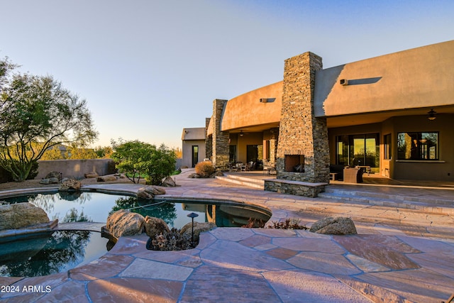 pool at dusk with ceiling fan, a patio area, and an outdoor stone fireplace