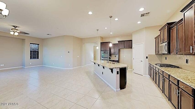 kitchen with appliances with stainless steel finishes, backsplash, light stone counters, ceiling fan, and a center island with sink