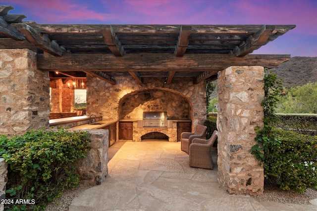 patio terrace at dusk with a pergola, a grill, exterior kitchen, sink, and an outdoor stone fireplace