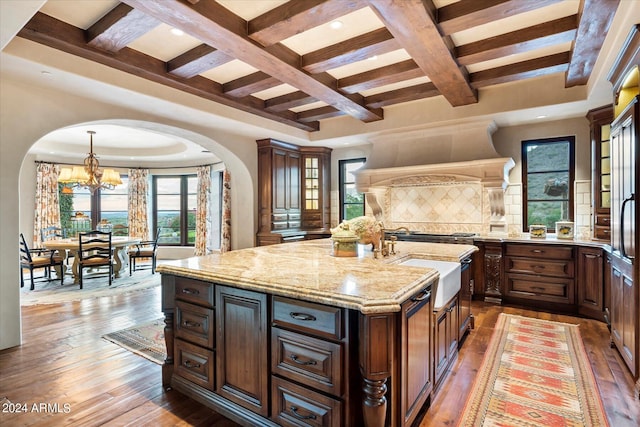 kitchen with coffered ceiling, beamed ceiling, light stone countertops, dark hardwood / wood-style flooring, and an island with sink