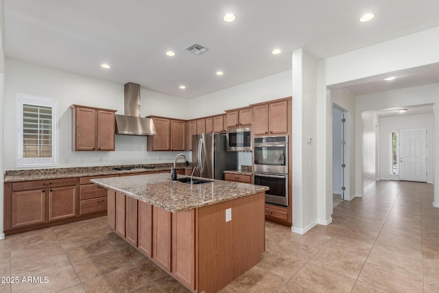 kitchen with wall chimney range hood, appliances with stainless steel finishes, a sink, an island with sink, and light stone countertops