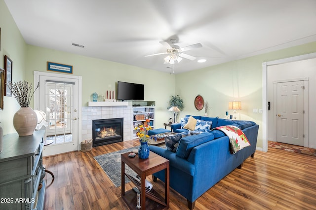 living area featuring a tiled fireplace, wood finished floors, baseboards, visible vents, and ceiling fan