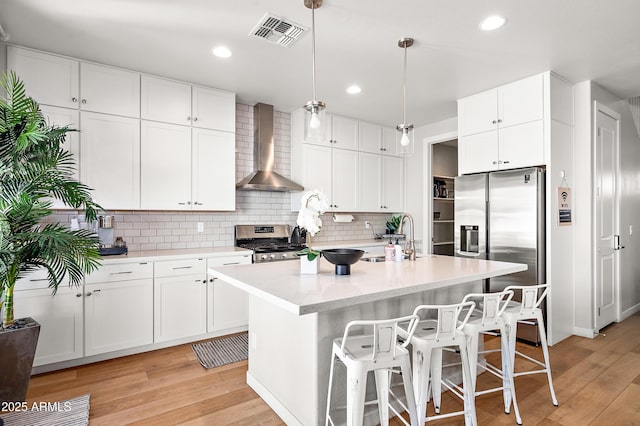 kitchen with decorative light fixtures, wall chimney range hood, a kitchen island with sink, stainless steel appliances, and white cabinets