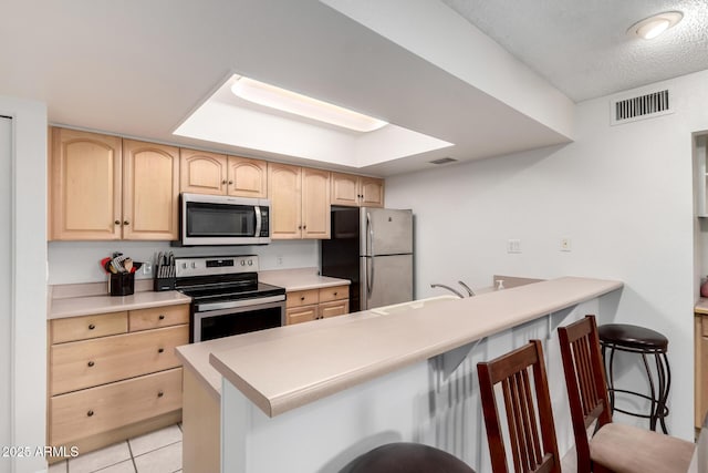 kitchen with light brown cabinetry, a breakfast bar area, light tile patterned floors, kitchen peninsula, and stainless steel appliances