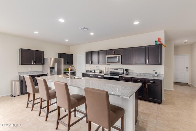 kitchen with sink, light stone counters, a center island with sink, appliances with stainless steel finishes, and a kitchen breakfast bar