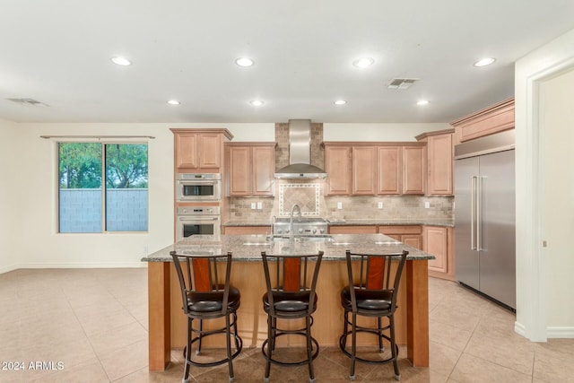 kitchen featuring wall chimney exhaust hood, light stone countertops, stainless steel appliances, and a kitchen island with sink