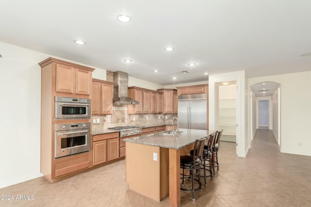 kitchen featuring a center island with sink, a kitchen breakfast bar, wall chimney exhaust hood, stone countertops, and stainless steel appliances