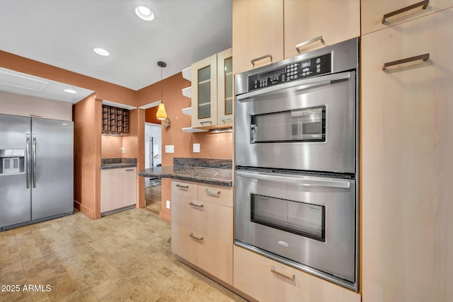 kitchen featuring stainless steel appliances, glass insert cabinets, light brown cabinets, decorative light fixtures, and dark stone countertops