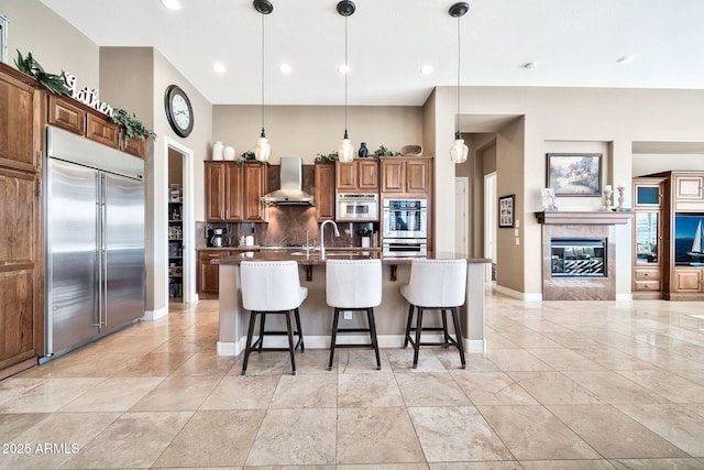 kitchen featuring pendant lighting, appliances with stainless steel finishes, wall chimney range hood, a kitchen island with sink, and a breakfast bar