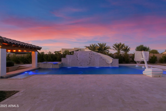 pool at dusk with an in ground hot tub, a patio, a water slide, and pool water feature