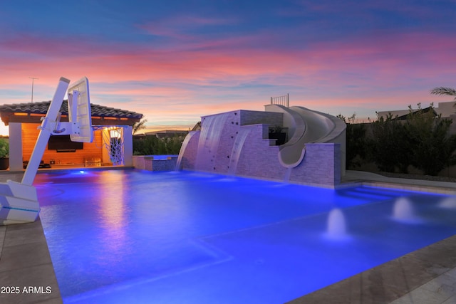 pool at dusk with pool water feature, a patio, and a water slide