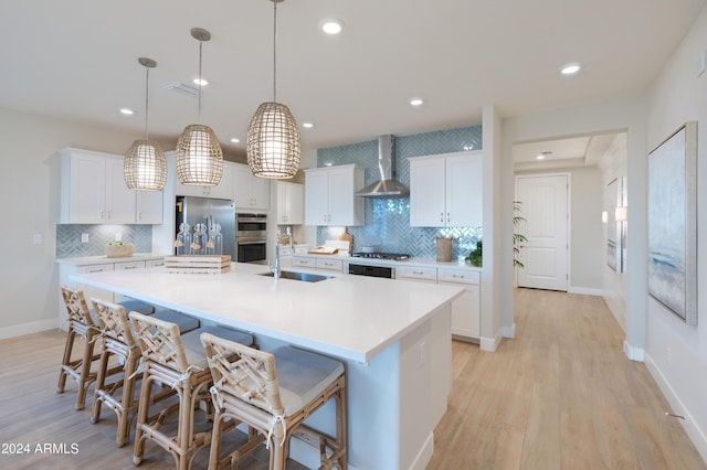 kitchen featuring a large island, wall chimney range hood, appliances with stainless steel finishes, white cabinets, and decorative light fixtures