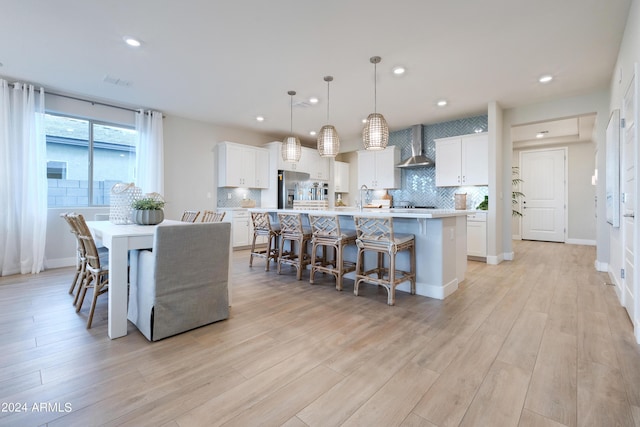 kitchen featuring stainless steel refrigerator with ice dispenser, wall chimney exhaust hood, white cabinetry, decorative light fixtures, and a center island with sink