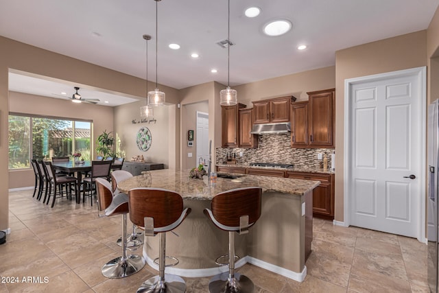 kitchen with light stone countertops, stainless steel gas cooktop, sink, decorative light fixtures, and an island with sink